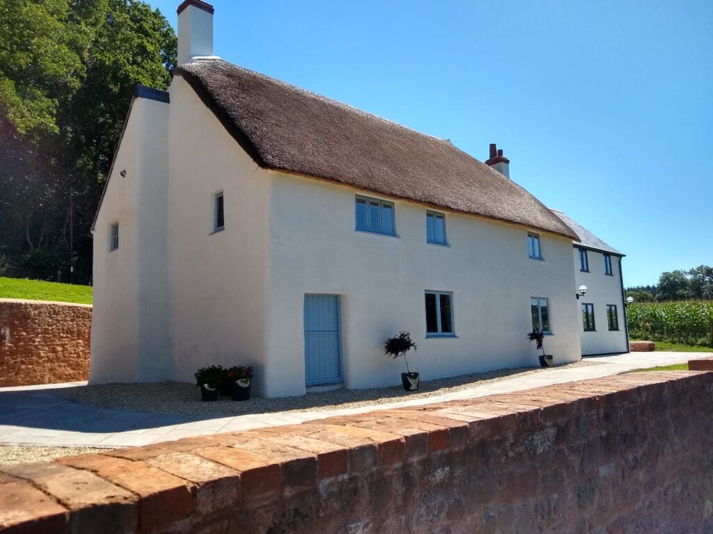 A white, two-story cottage with a thatched roof and blue-framed windows sits behind a low stone wall, showcasing charming house renovations amid greenery under a clear blue sky.