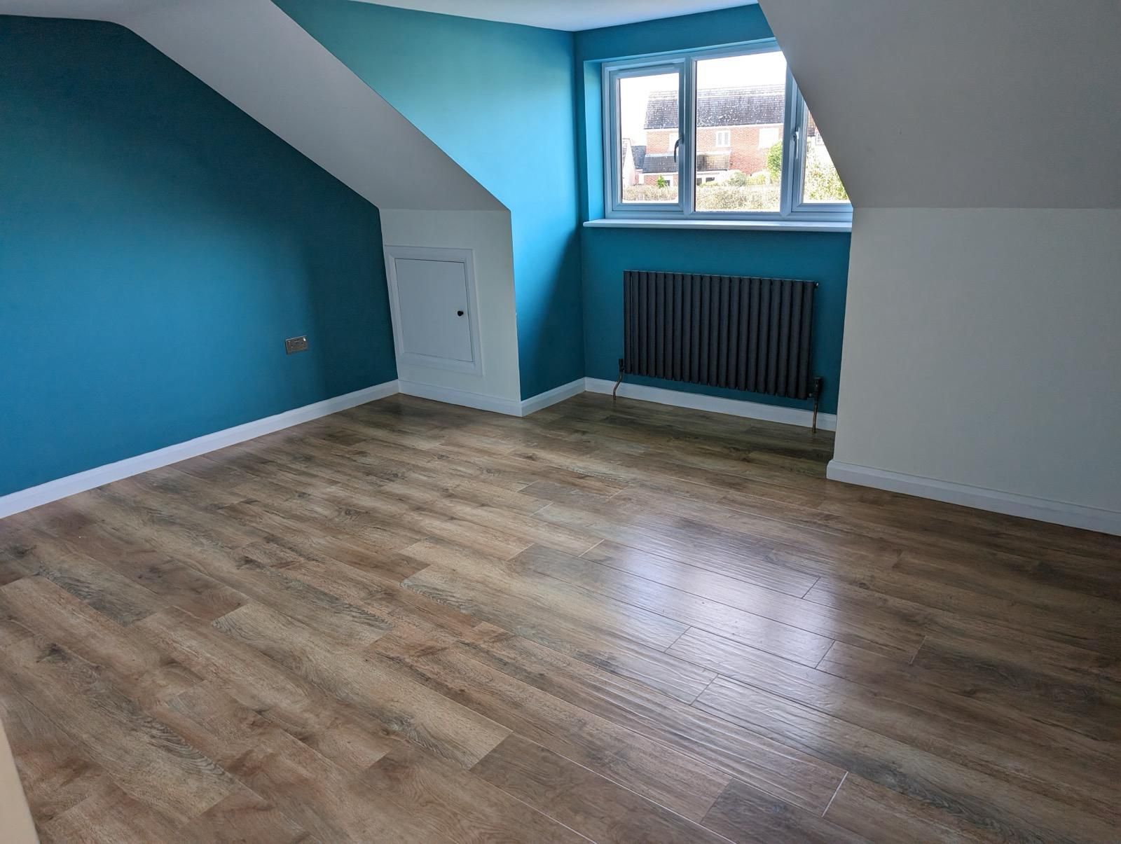 A bright, empty room with wooden flooring, teal and white walls—perfect for loft conversions. Features a small window with a black radiator beneath it and a small white door set into the sloped ceiling. Ideal for new build homes.