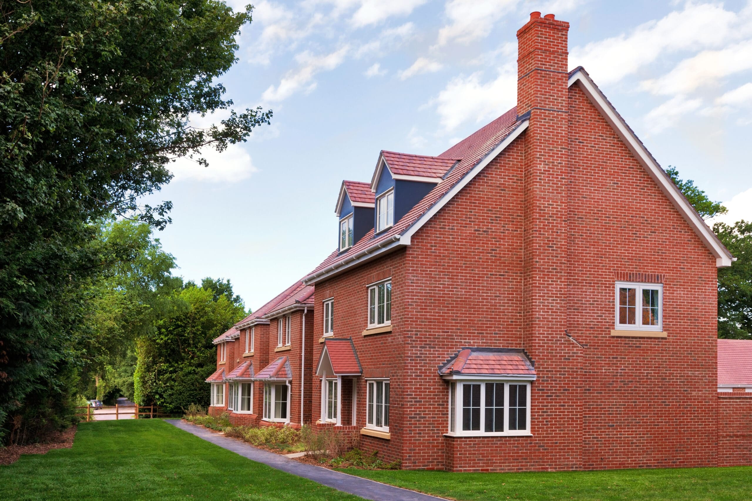 A large, modern red-brick new build home with white-framed windows and dormer windows on the roof, surrounded by green grass, trees, and a clear sky with scattered clouds.