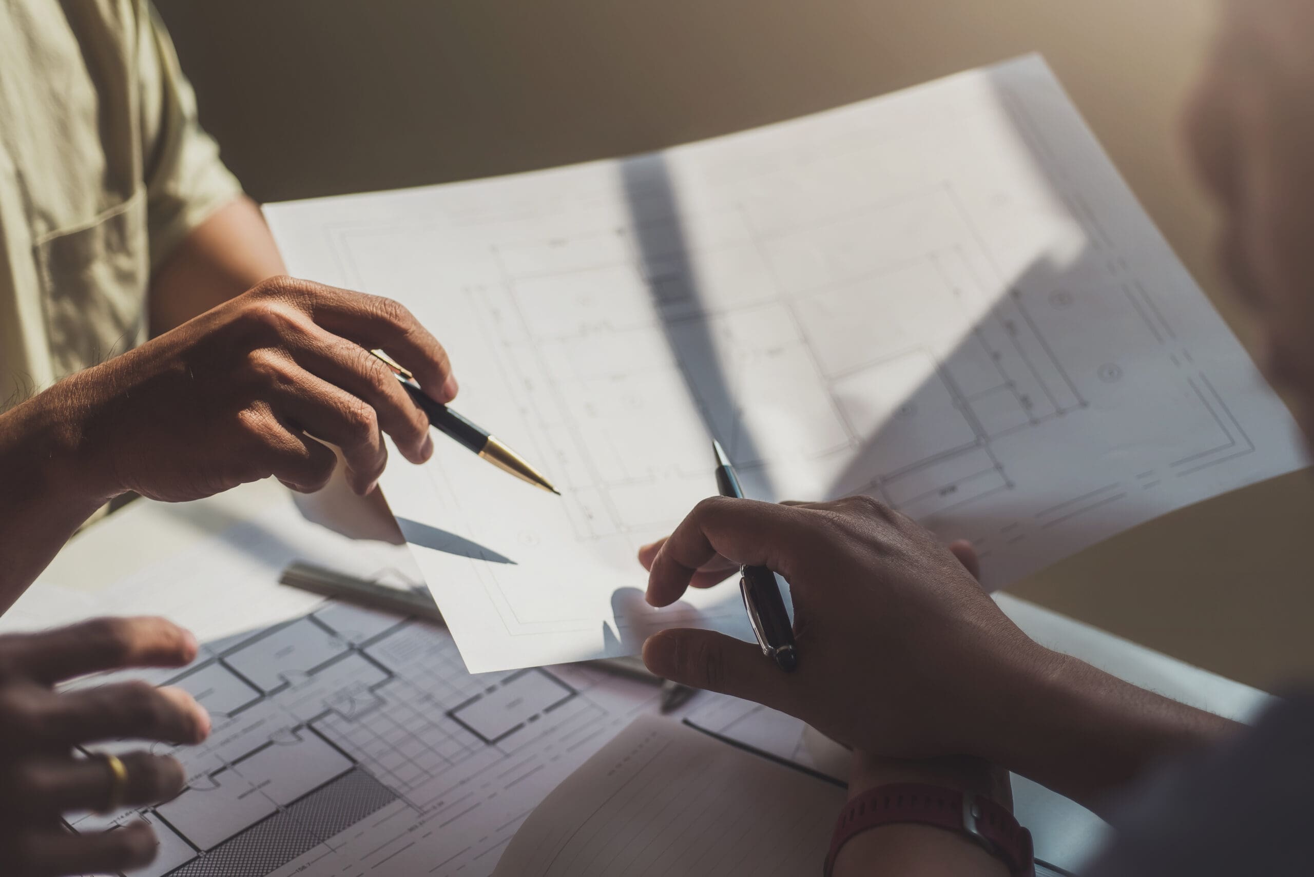Two people are reviewing and discussing architectural blueprints for a home extension at a table. One points to the plans with a pen, while the other writes on a paper, sunlight casting shadows over their builder documents.