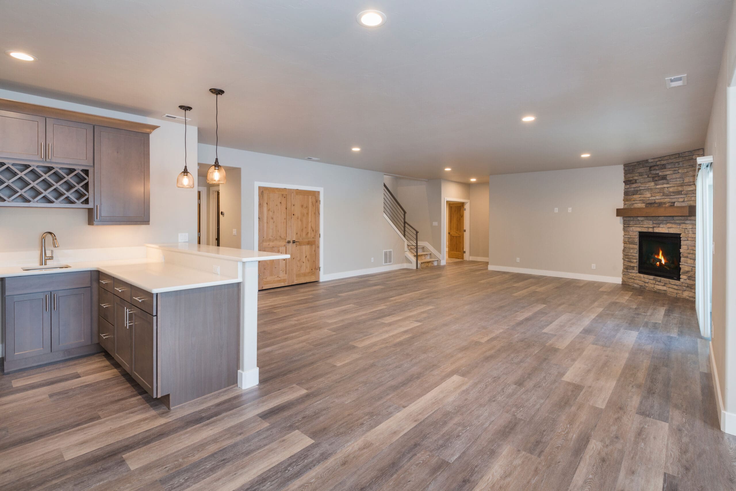 Spacious open-plan living area with wood-style flooring, modern kitchen with gray cabinets, pendant lights, a stone fireplace, and wooden double doors—ideal for house renovations or inspiration for new build homes. Staircase featured in the background.