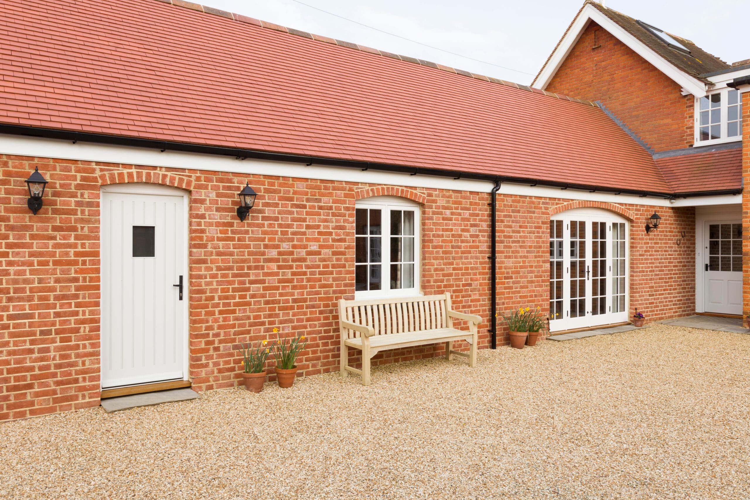 A red-brick house with a red-tiled roof, white doors and windows, potted plants, a wooden bench, and a gravel driveway in front—perfect for those considering home extensions or future house renovations.