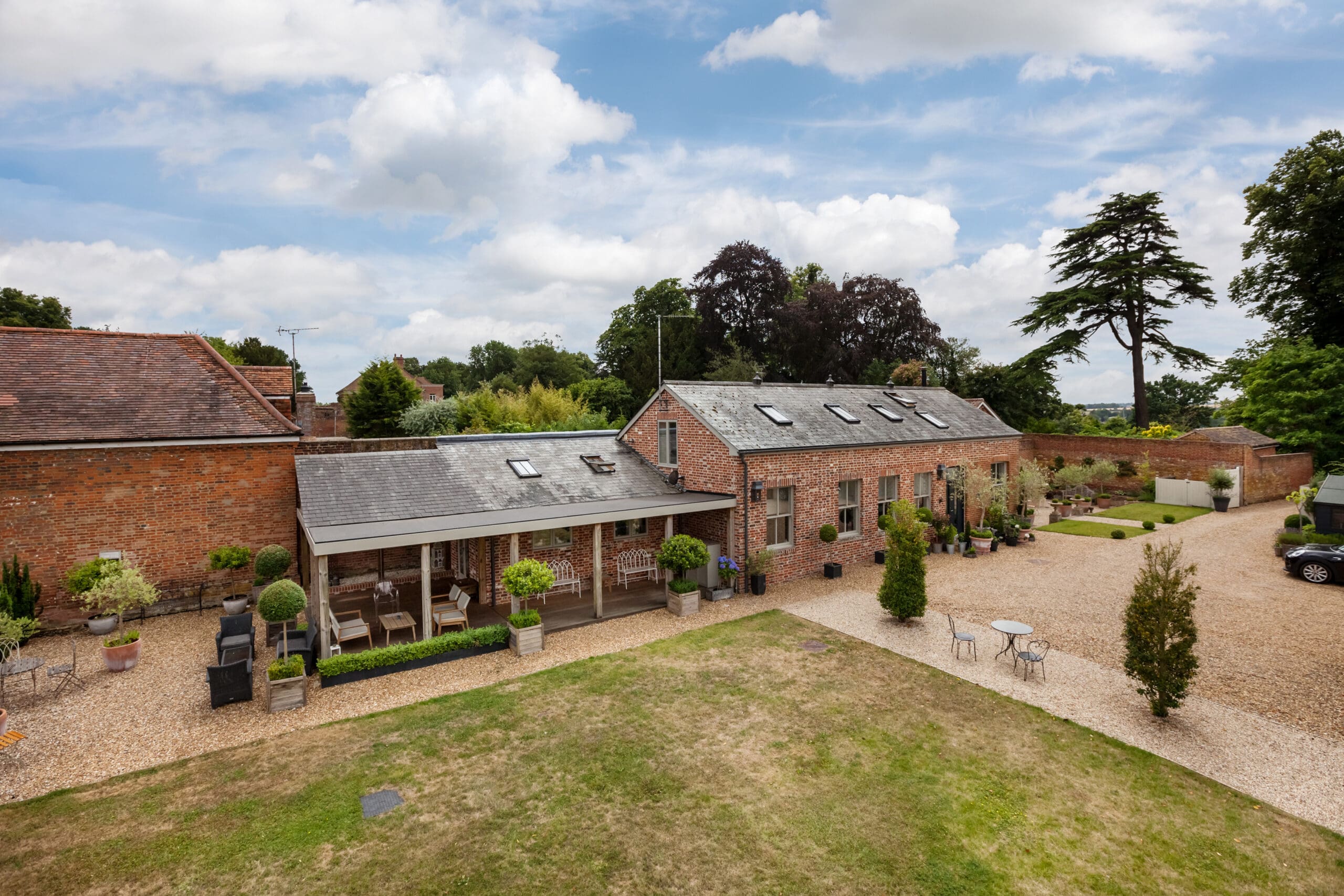 A large brick house with skylights, surrounded by gravel driveways, potted plants, and greenery, under a partly cloudy sky. Outdoor seating areas with tables and chairs are visible—ideal for those seeking home extensions or a skilled builder.