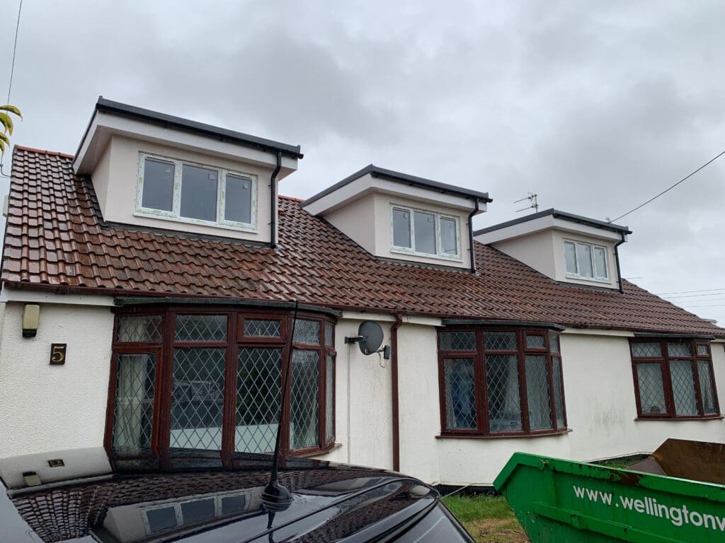 A white house with a tiled roof and three dormer windows. Brown-framed leaded windows, a black car in front, and a green skip on the right suggest house renovations are underway. The sky is cloudy.