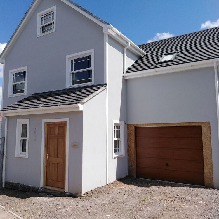 A modern two-story house with light gray walls, a wooden front door, white-framed windows, and a large wooden garage door—ideal for home extensions or loft conversions—set on a gravel driveway under a blue sky with scattered clouds.