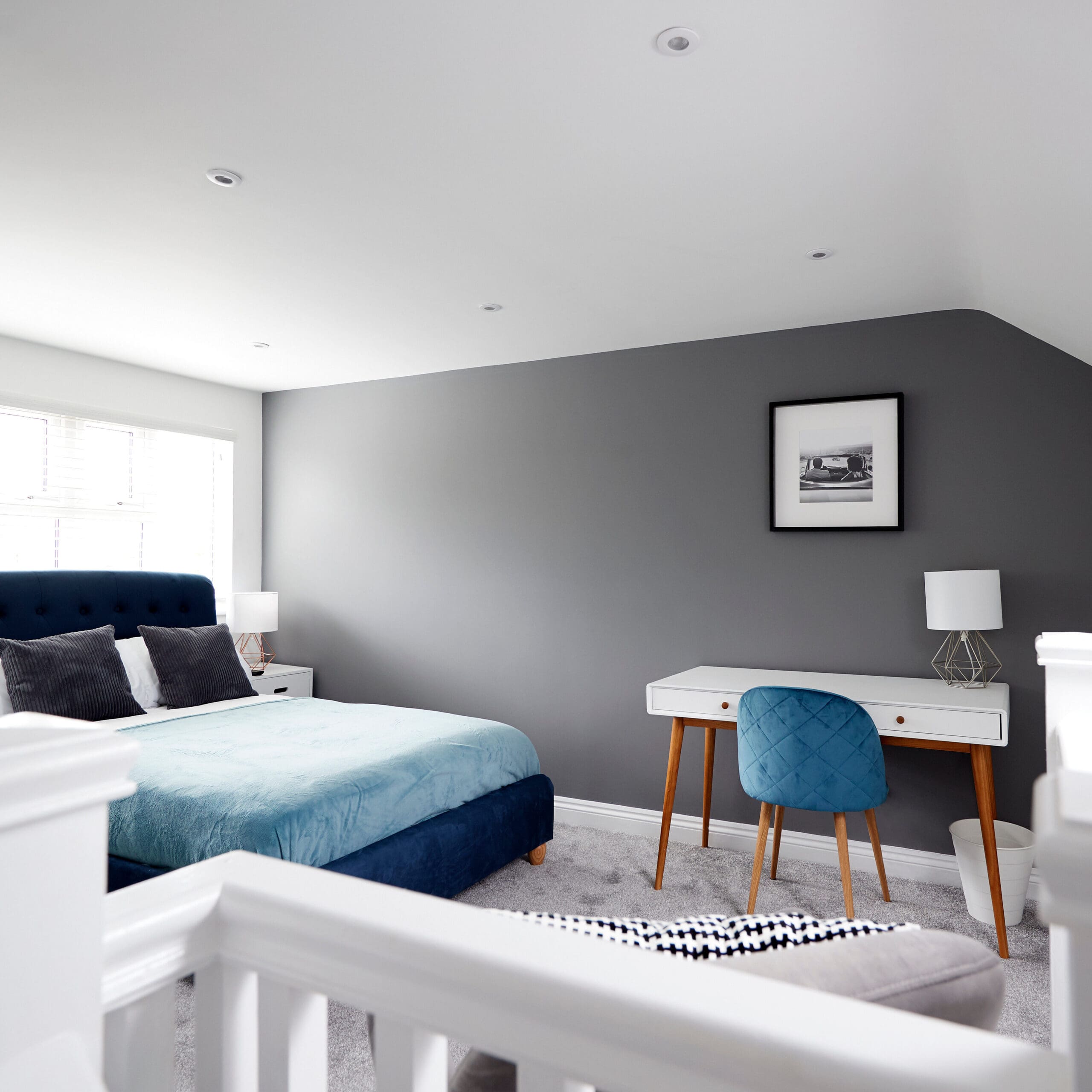 Modern bedroom with a teal bed, gray accent wall, white desk with a blue chair, lamps, and a framed picture. The room is bright with natural light coming from a window. White staircase railing is in the foreground.