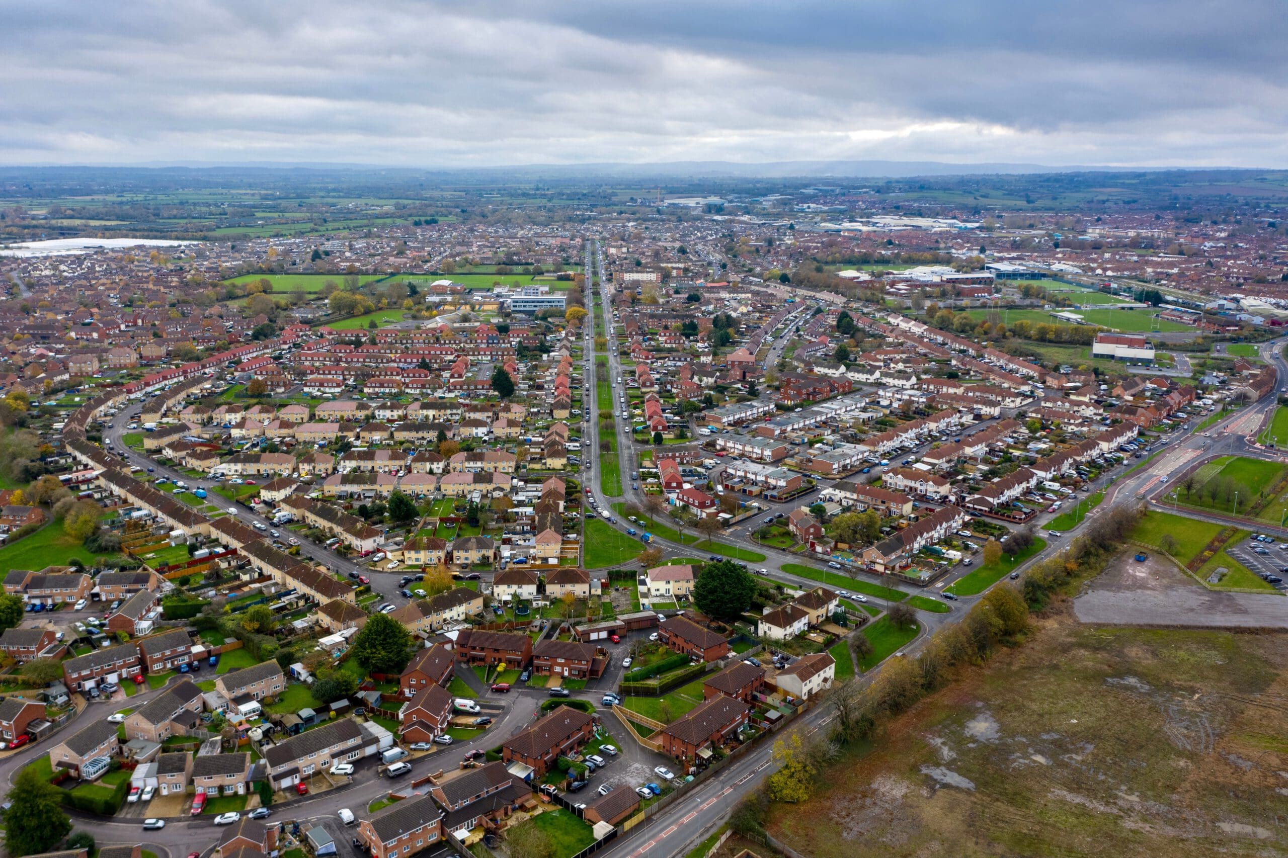 Aerial view of a suburban neighborhood with rows of houses, some featuring recent house renovations and loft conversions, intersecting roads, and green spaces under a cloudy sky. Fields and distant countryside stretch across the horizon.