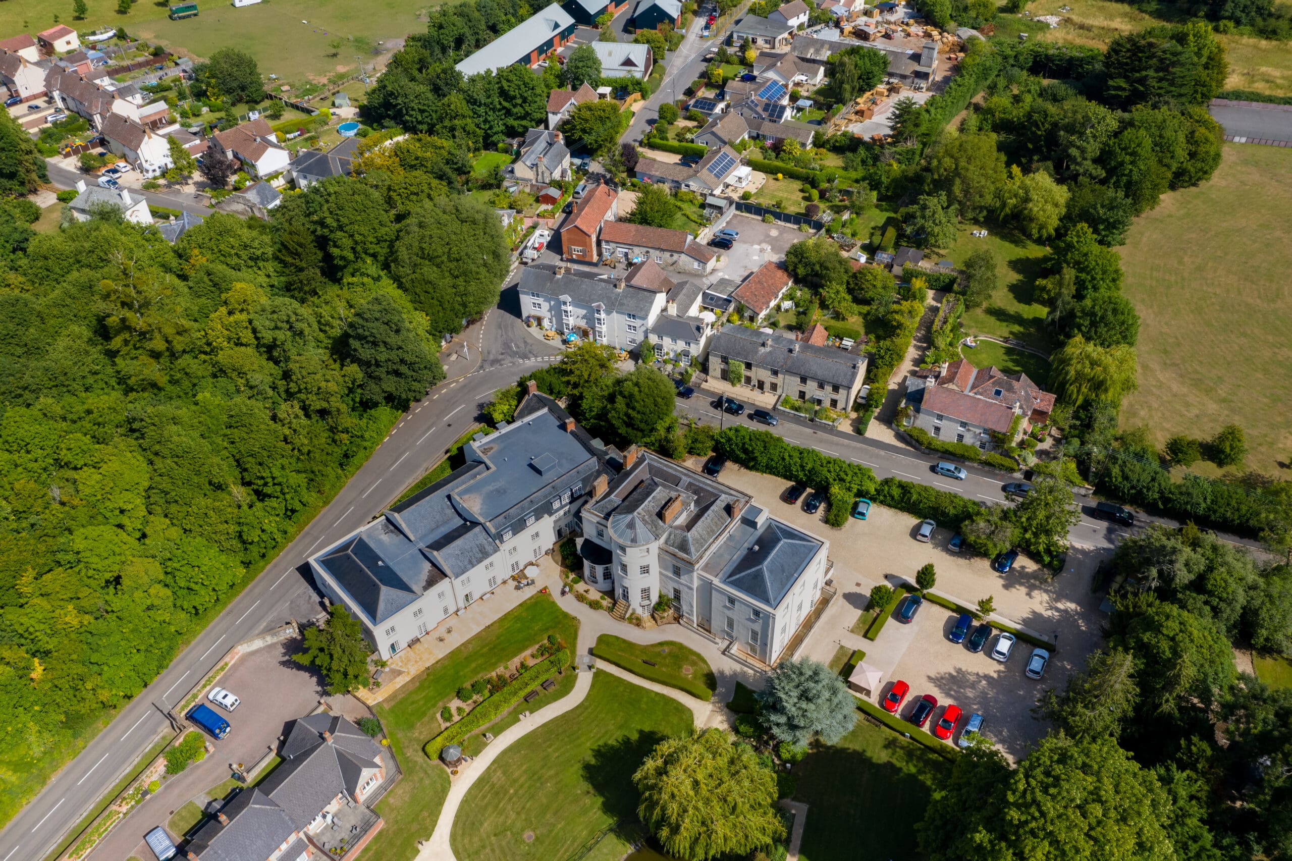 Aerial view of a large building with a curved driveway and parking lot, surrounded by trees and landscaped gardens, alongside newly built homes, residential houses, and roads in a suburban area.